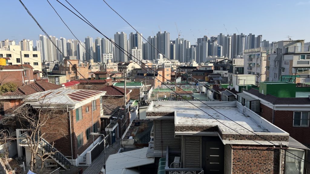 On the foreground, numerous low buildings, many with red bricks, some having a flat roof. In the background, a line of similar looking high apartment buildings.