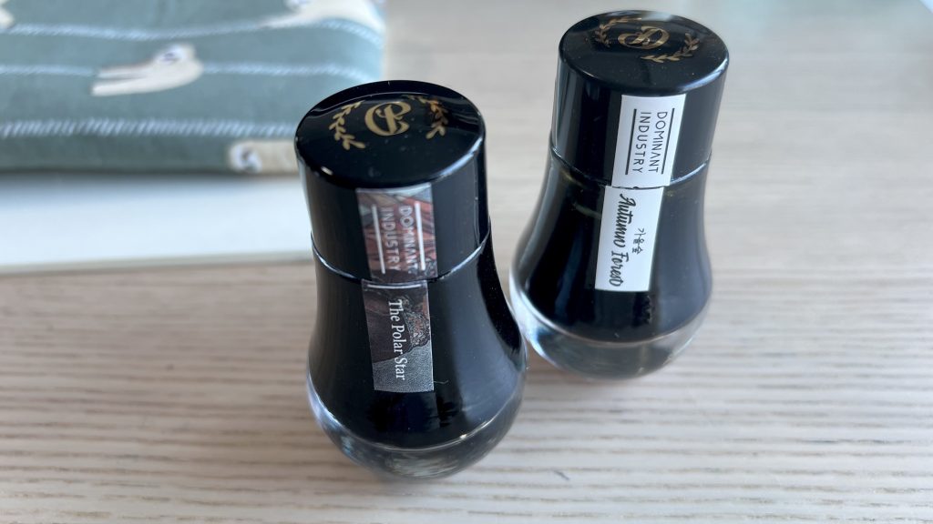 Two bottles of ink on a wooden table. A notebook and a pouch are blurred in the background. The two bottles are from the same brand, have an elegant curved body and display a label that says “Dominant Industry” and “The Polar Star” for the bottle on the left and “Autumn Forest” for the bottle on the right.
