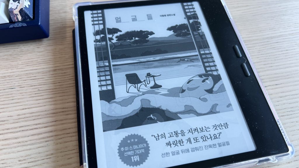 E-reader on a table displaying the cover of a book in Korean with a promotional wrapping band. The cover illustration shows masks in the foreground and a swimming pool with trees and hills in the background.