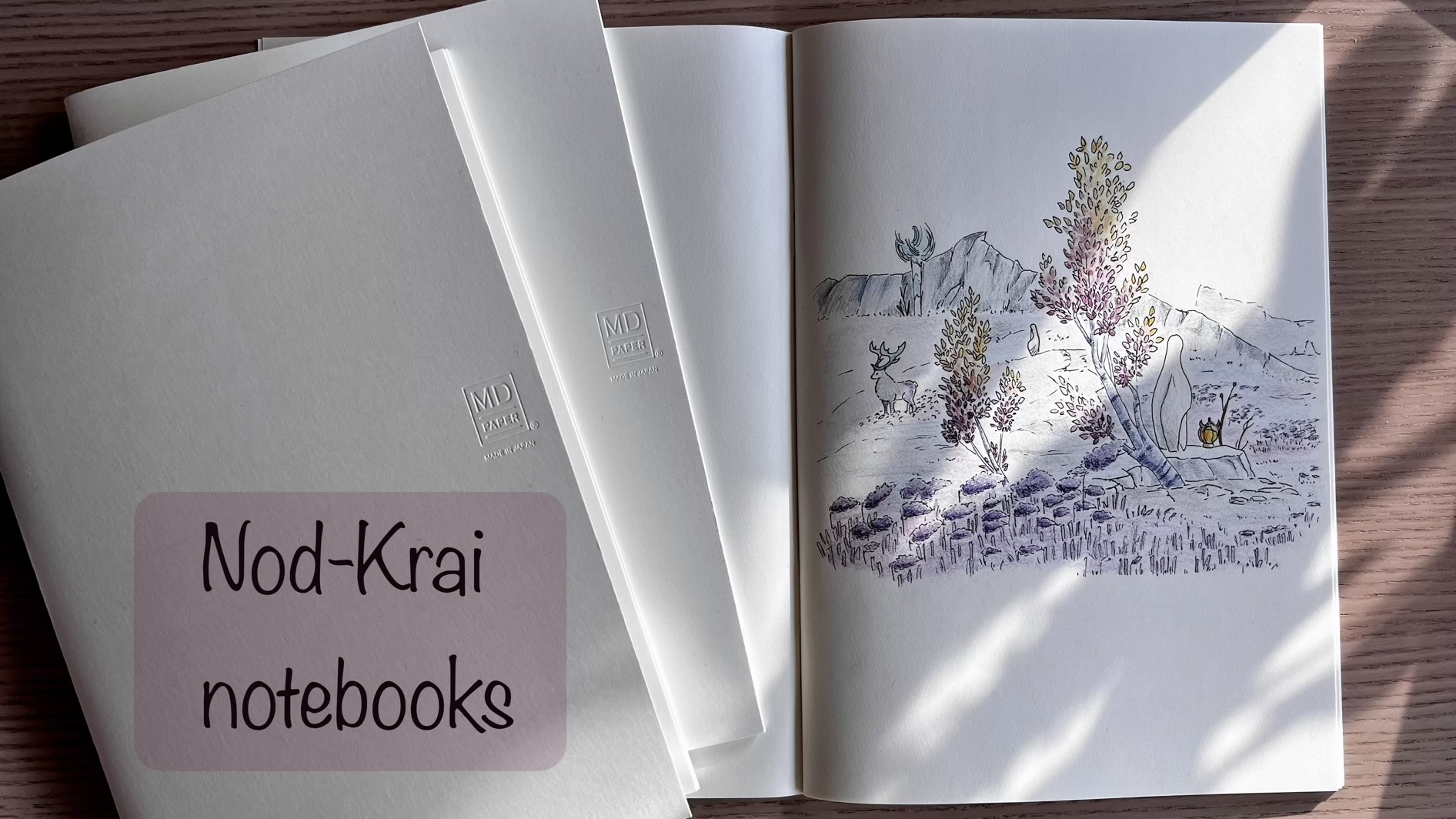 An opened notebook is displaying a drawing of a fantasy landscape with colours. Two white closed notebooks are resting beside it. The words “Nod-Krai notebooks” have been added on a pink background on the bottom left corner of the picture.